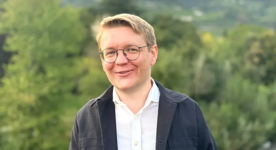 Ein Mann mit Brille lächelt in die Kamera. Er trägt ein weißes Hemd und eine dunkle Jacke. Im Hintergrund sind Bäume und Berge zu sehen. Er studiert Seelsorge.