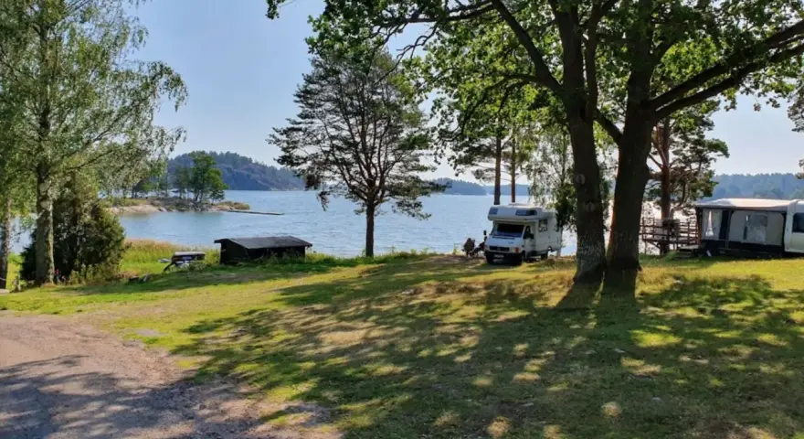 Solig dag vid vattnet på Kalvholmens camping. Husbilar och husvagnar står uppställda på gräsmattan nära sjön.