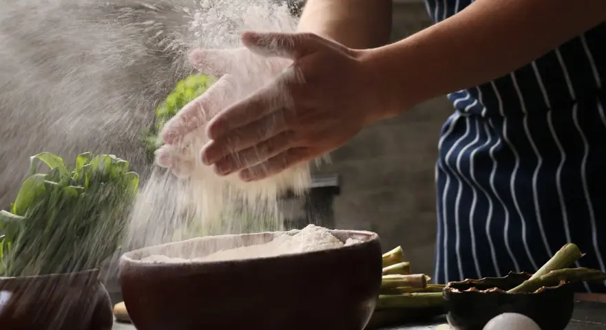 Hände verarbeiten Maniokmehl, das in der Luft schwebt. Daneben liegen Spargel und Eier, bereit für die Zubereitung.