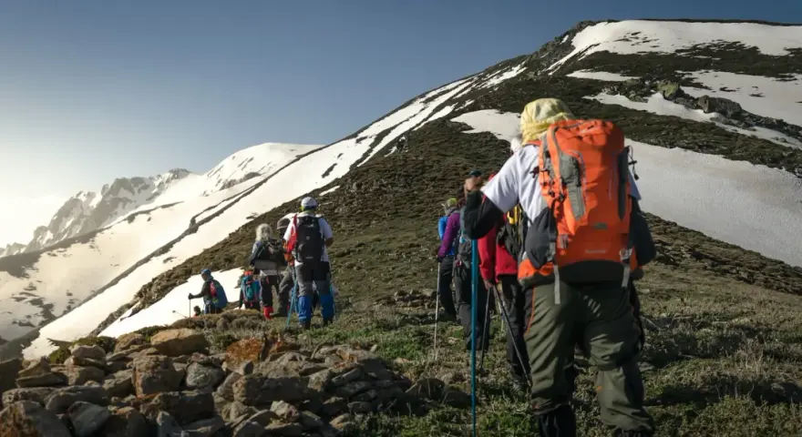 Un groupe de randonneurs progresse sur un sentier rocailleux, le soleil éclairant les montagnes enneigées. Ils semblent déterminés à savoir son altitude.