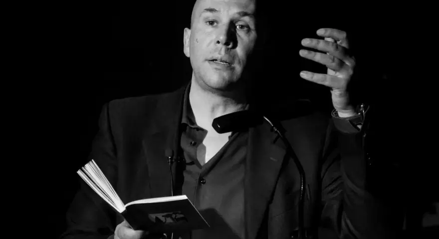 Un uomo calvo, narratore protagonista, legge da un libro aperto, gesticolando con la mano.