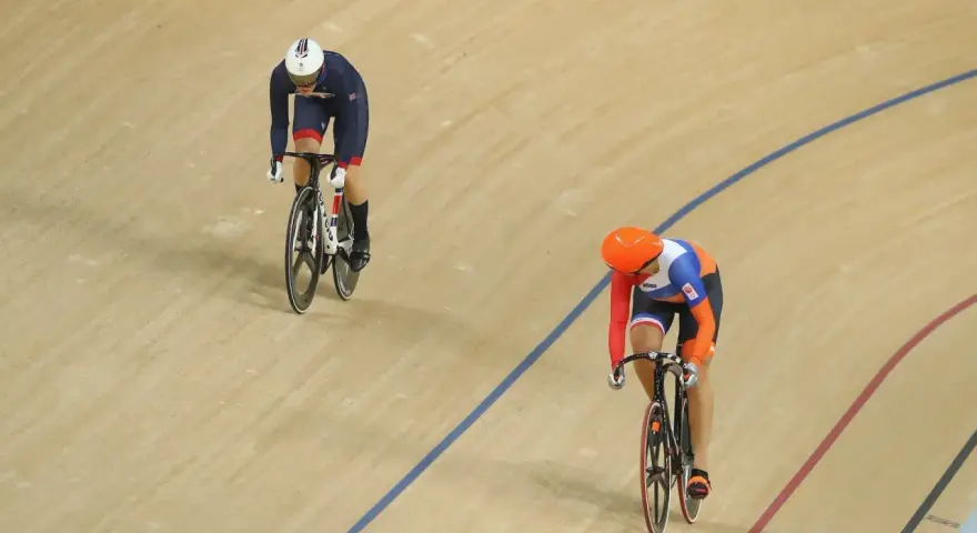 Two cyclists compete in a track sprint. One rider in a blue and white uniform leads, while another in orange and blue follows closely.