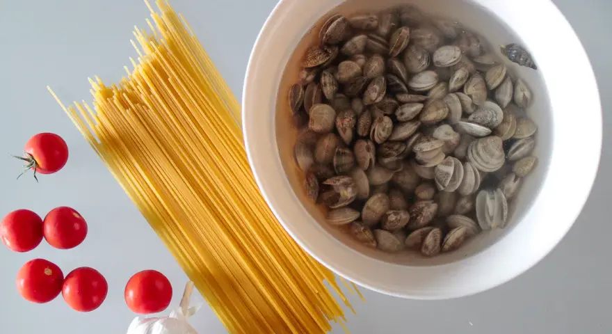 Składniki na pyszny makaron z małżami: spaghetti, pomidorki, czosnek i świeże małże w misce.
