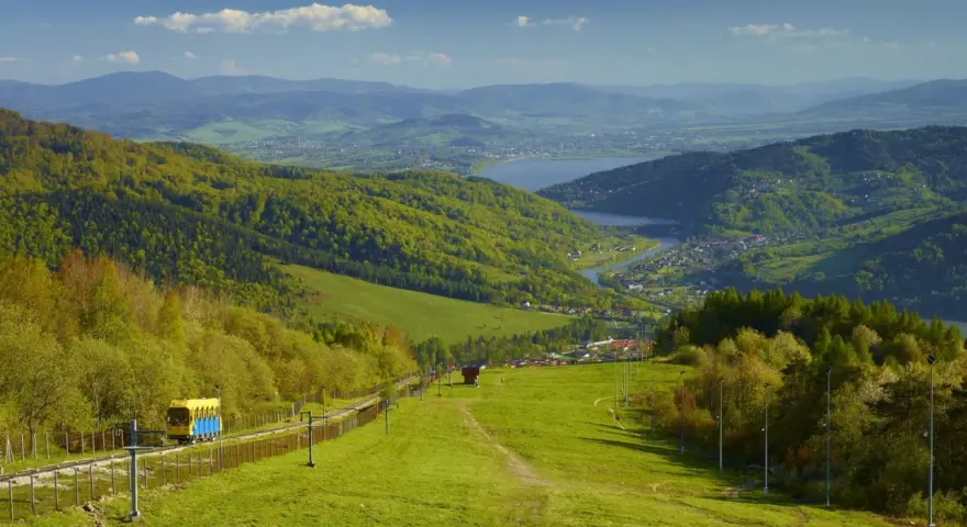 Widok na zielone **beskid mały szczyty** z kolejką linową i jeziorem w oddali.