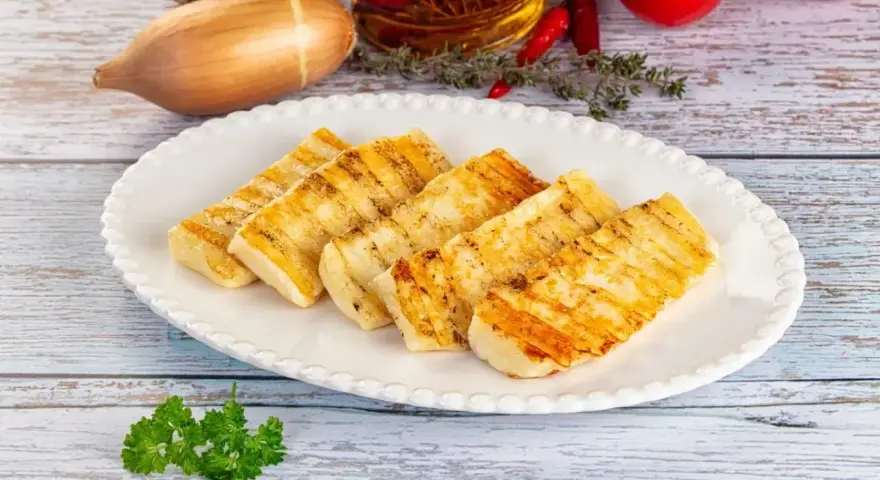 Grillowany ser halloumi na białym talerzu, z dodatkami jak czosnek, cebula i pomidory w tle.