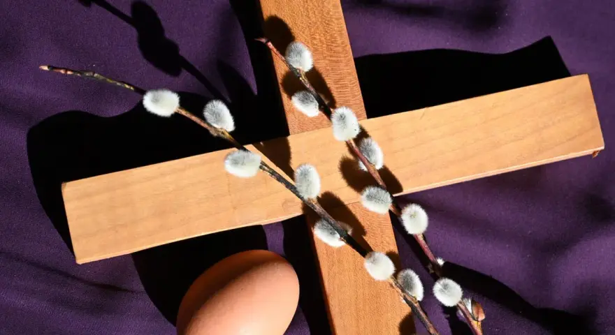 Holzkreuz mit Weidenkätzchen und Ei auf lila Stoff, ein Symbol für christliche Ostern.