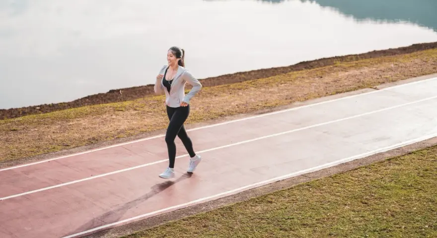 Une femme souriante profite d'une marche rapide sur une piste de course, au bord d'un lac paisible.