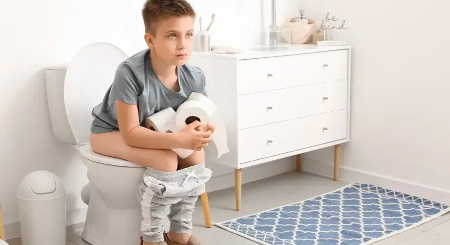 Chłopiec siedzi na toalecie, trzymając rolki papieru toaletowego. Wygląda na chorego, co może sugerować wymioty i biegunka u dziecka.