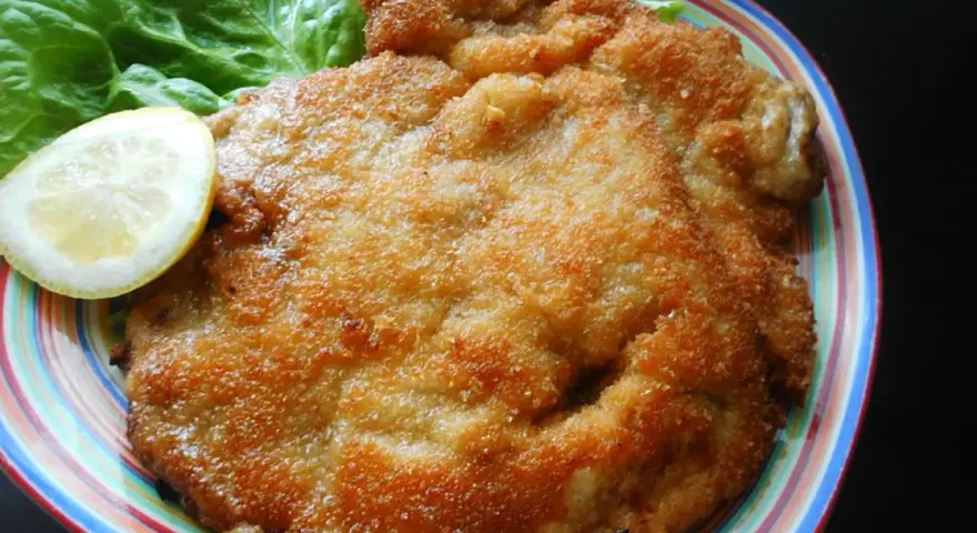 Złocisty kotlet schabowy Magdy Gessler, podany z liściem sałaty i plasterkiem cytryny na kolorowym talerzu.