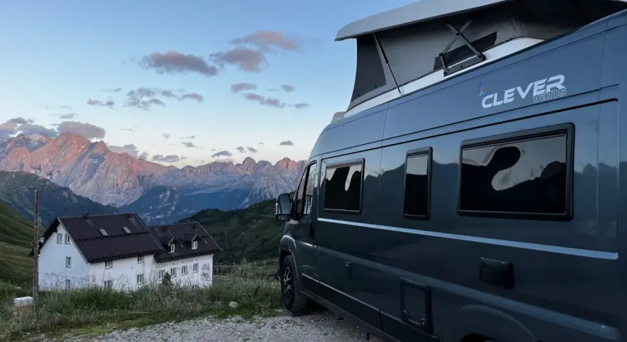 En mörkgrå husbil med pop-up tak, märkt "Clever vans", står parkerad vid en bergig stuga med Alperna i bakgrunden.