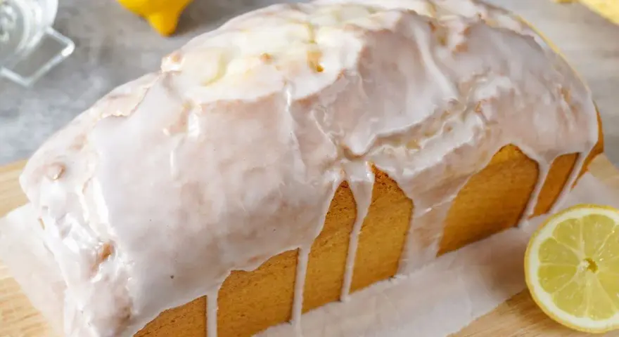 Saftiger zitronenkuchen in Kastenform, überzogen mit süßem Zuckerguss. Ein Zitronenstück liegt daneben.