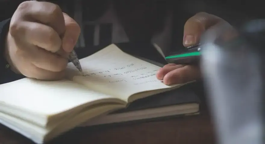 Dłoń pisze piórem w notatniku, przygotowując się do napisania rozprawki.