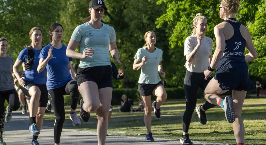 En grupp kvinnor tränar löpning utomhus. De springer i ett parkliknande område, redo att springa 1 km tid.