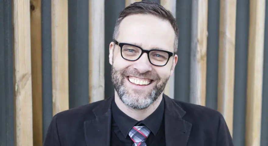 A smiling man with glasses and a beard, wearing a suit and tie, stands in front of a wooden slatted background.