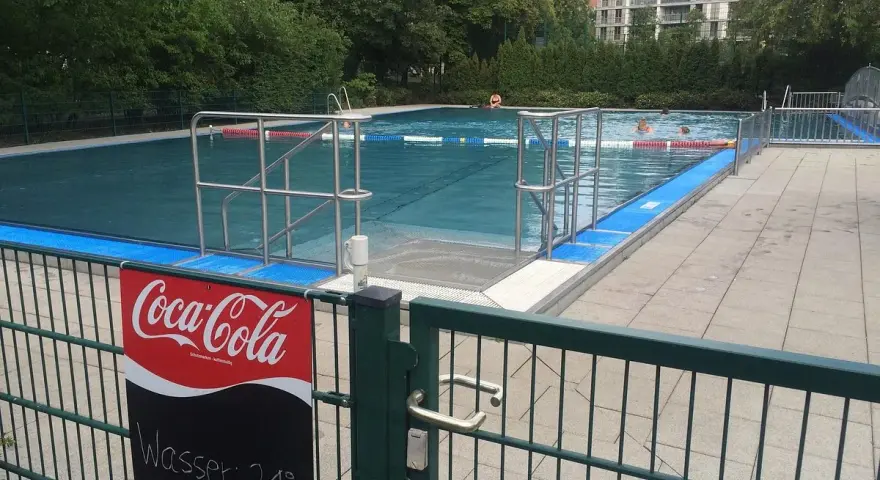 Erfrischung im Monbijou Bad: Wasser 21°C. Menschen schwimmen im blauen Becken, umgeben von Bäumen und einem modernen Gebäude.