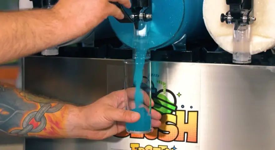 Ein Arm mit einem Ketten-Tattoo füllt ein Glas mit blauem Slush Eis aus einem Automaten.
