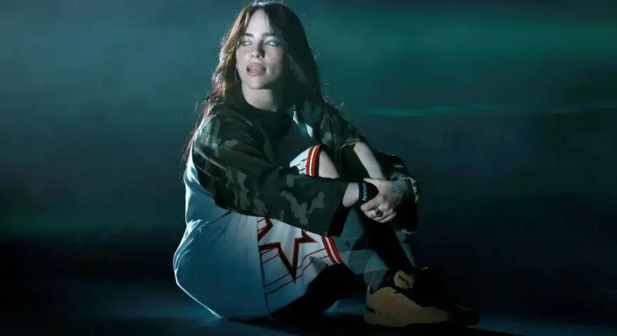 Billie Eilish w ciemnym pomieszczeniu, siedzi w pozie zamyślenia. Jej stylizacja nawiązuje do estetyki jej albumu.