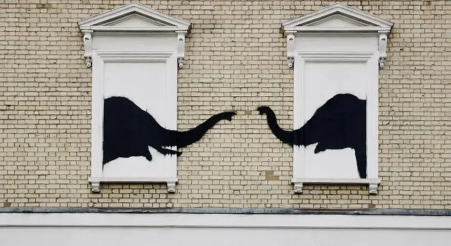 Œuvre Banksy : deux éléphants noirs se touchent la trompe à travers deux fenêtres sur un mur de briques.