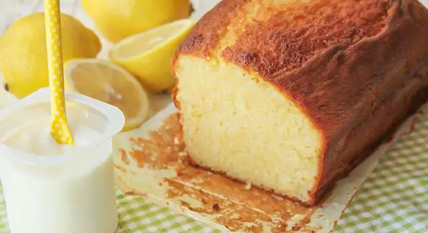 Bizcocho de yogur casero, dorado y esponjoso, acompañado de limones frescos y un vasito de yogur.