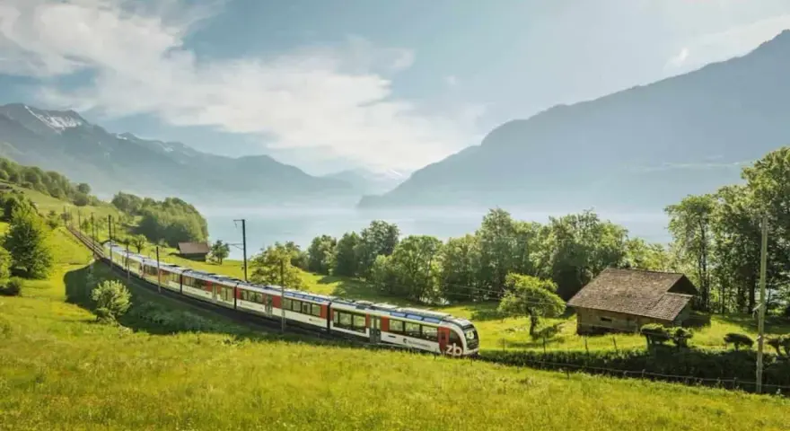 A train travels through a lush green valley, with a lake and mountains in the background. This scenic route from Lucerne to Montreux offers breathtaking views.