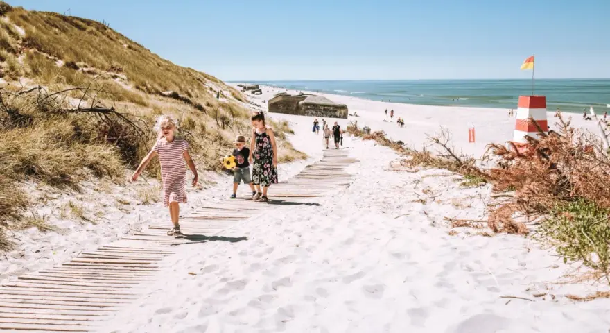 Familj promenerar på en strand med vit sand i Danmark. En livräddartorn och bunkrar syns i bakgrunden.