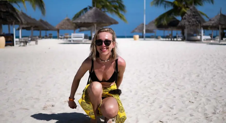 Uśmiechnięta blondynka w bikini i żółtej spódniczce kuca na piaszczystej plaży na Zanzibarze. W tle palmy i słomiane parasole.