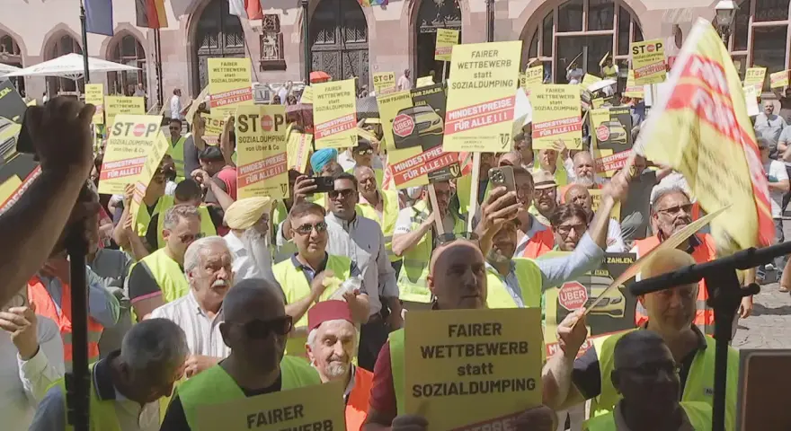 Demonstranten mit gelben Schildern fordern "Fairer Wettbewerb statt Sozialdumping" und "Mindestpreise für alle". Ein **Taxi-Streik** ist im Gange.