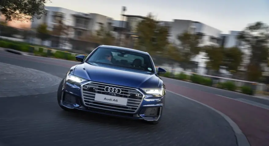 Niebieskie Audi A6 C8 w ruchu, z rozmazanym tłem budynków i drzew. Nawet przy potencjalnych usterkach, wygląda imponująco.