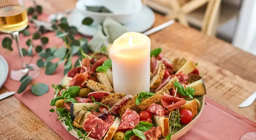 Antipasti selber machen: Eine festliche Platte mit Wurst, Käse, Brot, Oliven und Tomaten, umgeben von grünen Zweigen, mit einer Kerze in der Mitte.