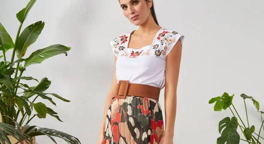 Mujer con top blanco bordado y falda plisada de estampado abstracto. Un cinturón marrón ancho realza la cintura.