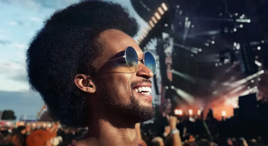 Homme souriant à un concert, portant des lunettes de soleil et un bouchon d'oreille pour protéger son audition.