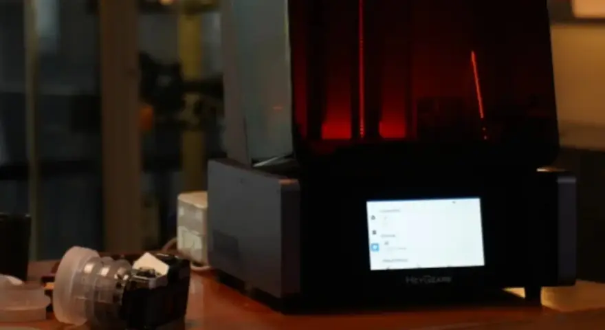 A HeyGears resin printer with a red interior light. Beside it, a camera lens. This setup hints at the process of how long to cure resin prints.