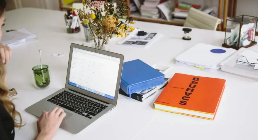 Escritorio con portátil, libros y flores. Un espacio de trabajo que parece una copistería por la cantidad de documentos y la organización.