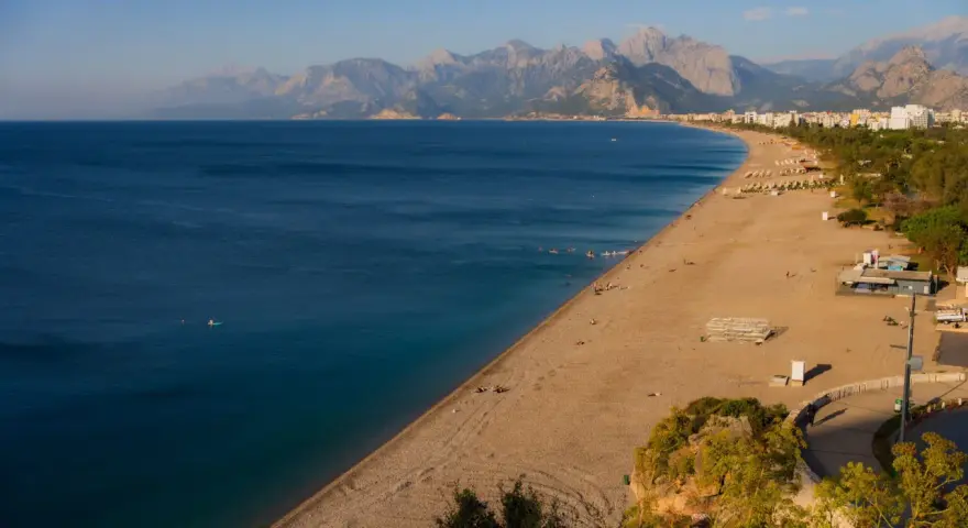 Słoneczna plaża na riwierze tureckiej z turkusowym morzem i górami w tle. Kilka osób pływa, inni odpoczywają na piasku.