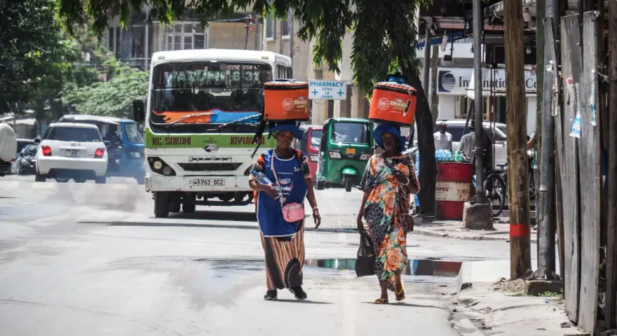 Dwie kobiety niosą na głowach pomarańczowe lodówki w upalny dzień w Dar es Salaam. W tle autobus i ruchliwa ulica.