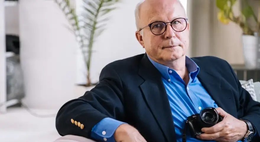 Tomaszewski fotograf w okularach, w granatowej marynarce i niebieskiej koszuli, trzyma aparat.