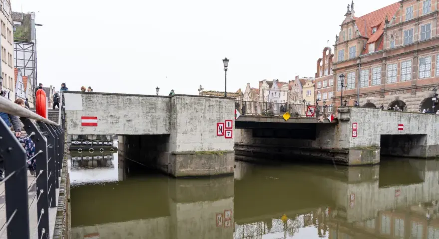 Nad kanałem w Gdańsku wznosi się betonowy zielony most, ozdobiony czerwono-białymi znakami. Ludzie spacerują po jego balustradzie.