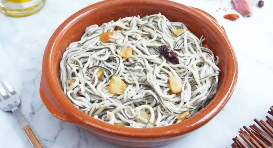 Un cuenco de barro con gulas al ajillo, adornado con láminas de ajo dorado y un chile seco.