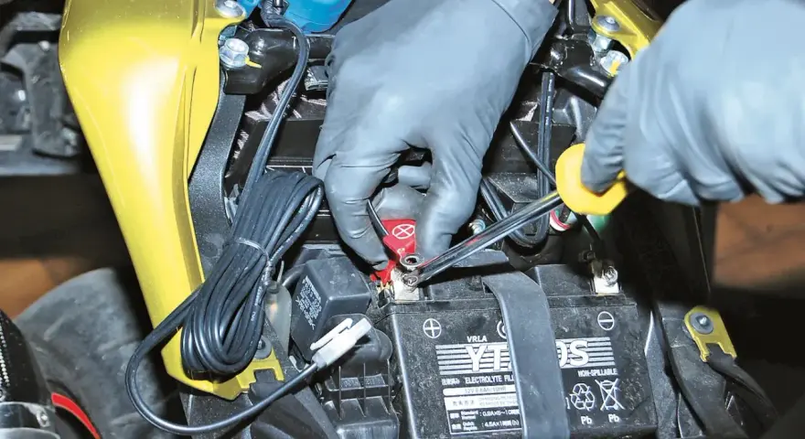 Mani guantate usano una chiave per staccare la batteria moto, scollegando il cavo rosso.