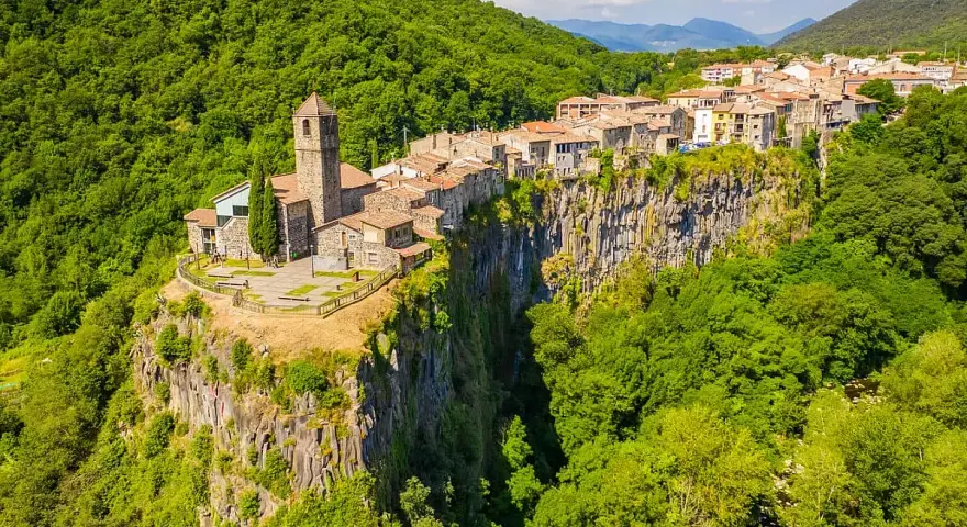 Un borgo medievale, Castellfollit de la Roca, aggrappato a una scogliera basaltica, circondato da lussureggianti foreste verdi.