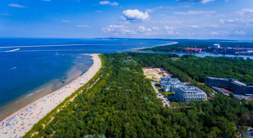 Widok z lotu ptaka na najszerszą plażę w Polsce, pełną wypoczywających ludzi, otoczoną lasem i nowoczesnymi budynkami.