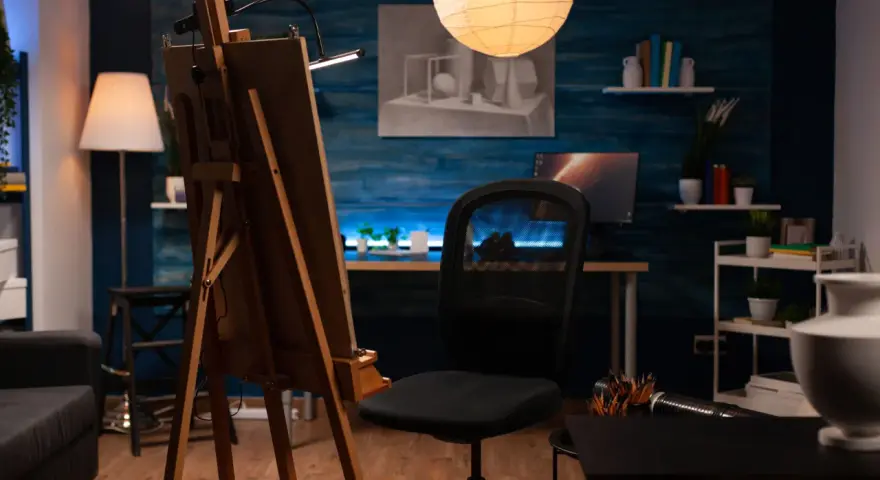 Un estudio de pintura en casa con caballete, silla de oficina, lámpara de pie y un escritorio con ordenador.