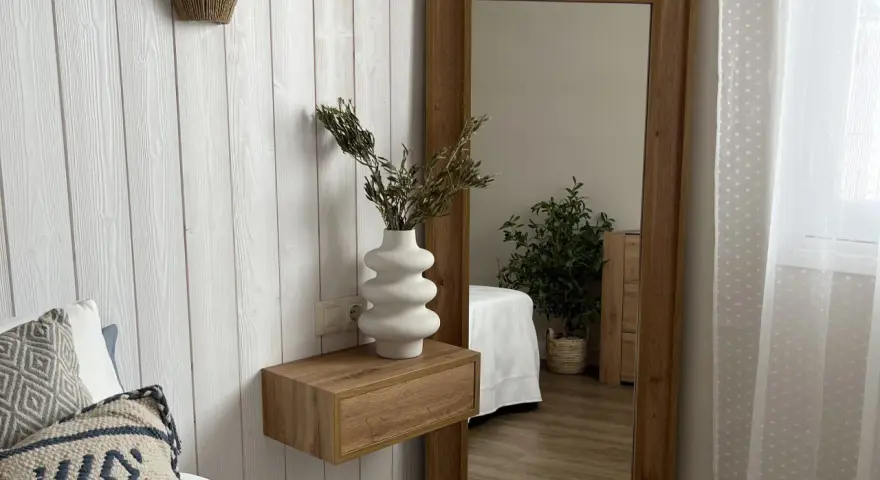 Estantería flotante de madera con jarrón blanco y ramas secas, ideal para ideas para decorar rincones pequeños. Reflejo de planta y cama en espejo grande.