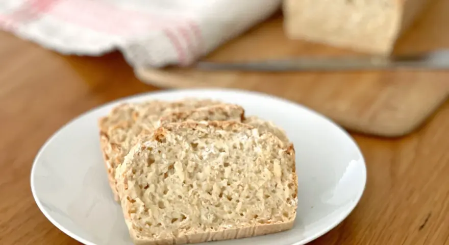 Skivor av hembakat bröd på en vit tallrik, perfekt för snabb bakning. En bit bröd ligger på en skärbräda i bakgrunden.