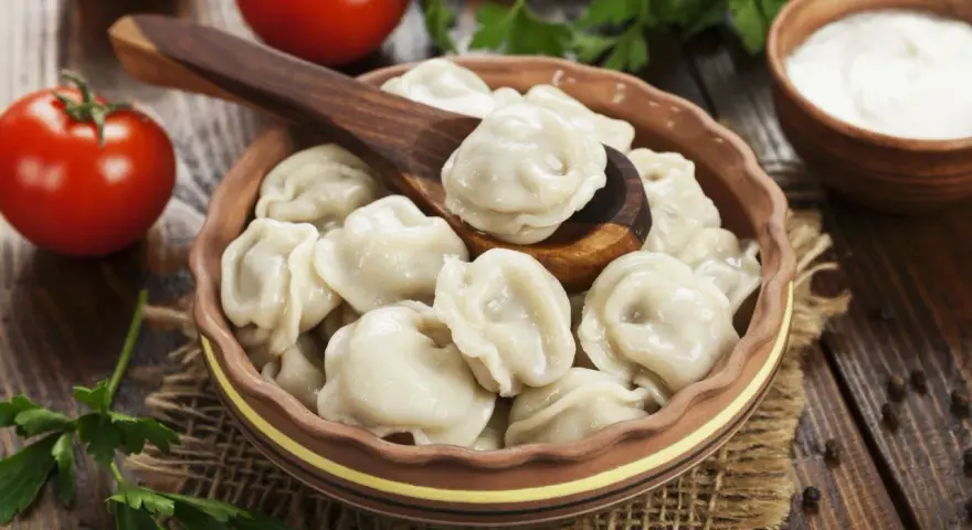 Pielmieni co to? Pyszne, gotowane pierożki w glinianej misce, z drewnianą łyżką, pomidorami i śmietaną w tle.