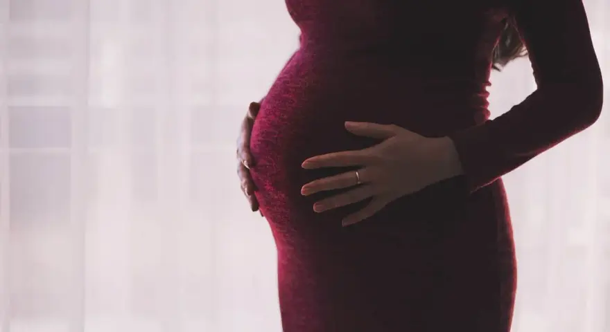 Kobieta w ciąży, z dłońmi na brzuchu, w ciemnoczerwonej sukience. Myśli o zasiłku macierzyńskim po ustaniu zatrudnienia.