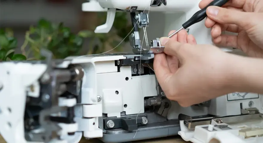 Dłonie regulują maszynę do szycia, używając śrubokręta. Widać fragmenty mechanizmu i nitki, a także pasek napędowy.
