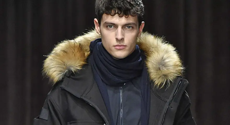 Modello con giacca Hugo marca con cappuccio bordato di pelliccia, sciarpa blu scuro, sguardo intenso.