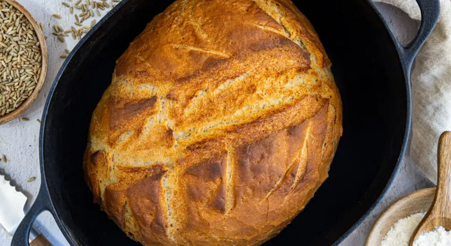 Ein goldbraunes Brot mit knuspriger Kruste liegt in einem Gusseisentopf. Dieses Brot kommt nicht aus dem Topf, sondern wurde darin gebacken.