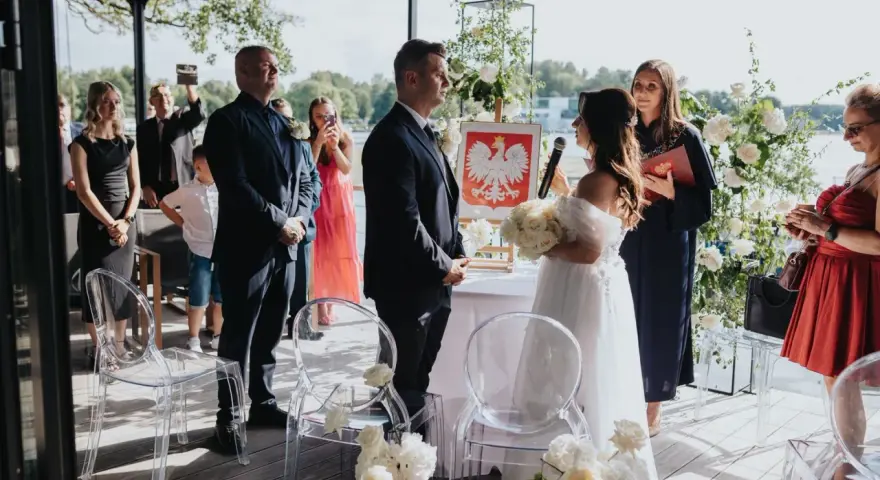 Uroczyste wesele w ogrodzie. Para młoda stoi przed urzędniczką, goście obserwują ceremonię.